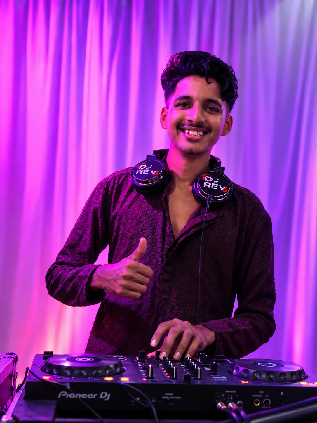 DJ Rev smiling with a thumbs up at a Pioneer controller against purple drapes