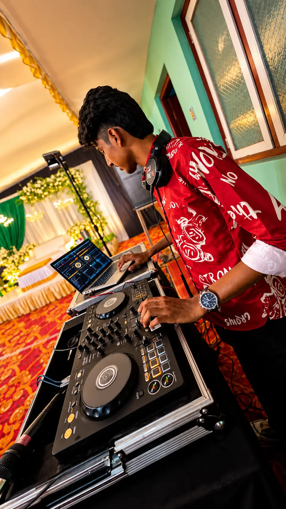 DJ Rev in a red printed shirt mixing beside a decorated wedding stage